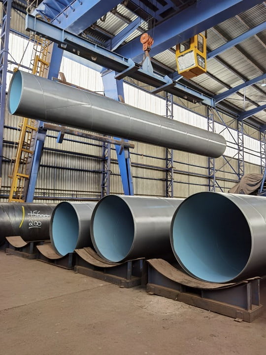 Workshop view of fabricated and lined large-diameter pipe spools under gantry crane